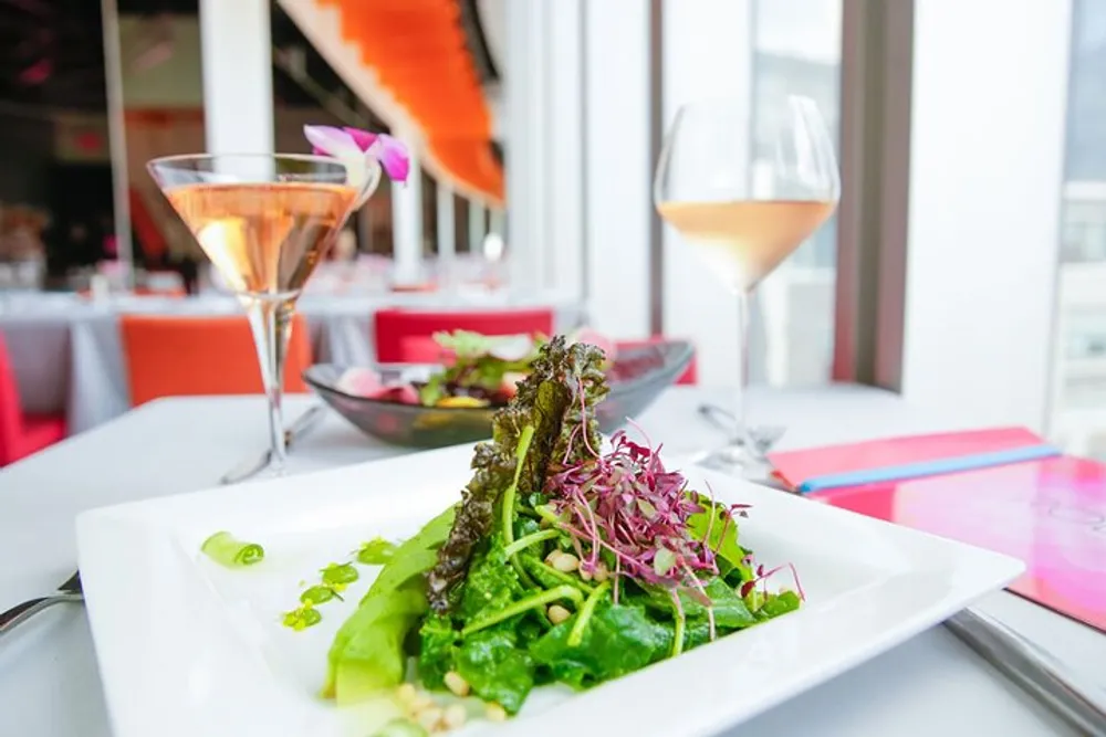 A fresh leafy green salad is presented on a white square plate in an elegant dining setting with glasses of ros wine in the background