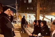 A group of people is sitting and standing on a city sidewalk at night, engaging in conversation, near a building labeled 