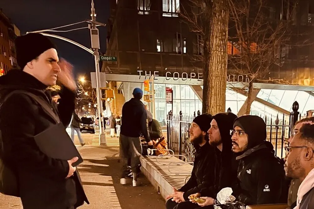 A group of people is sitting and standing on a city sidewalk at night engaging in conversation near a building labeled THE COOPER UNION