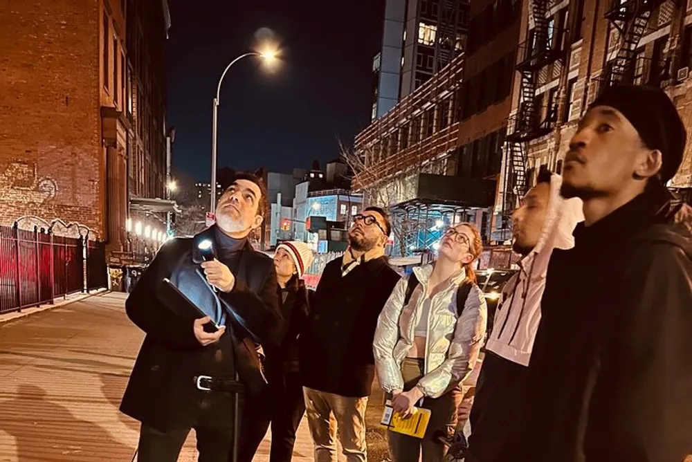 A group of people look upwards with interest and curiosity at night on an urban street possibly observing or listening to a tour guide