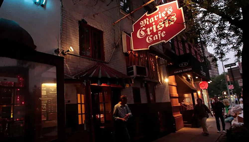 A person stands by the entrance of Maries Crisis Caf with a lit-up sign overhead as dusk falls on a busy street