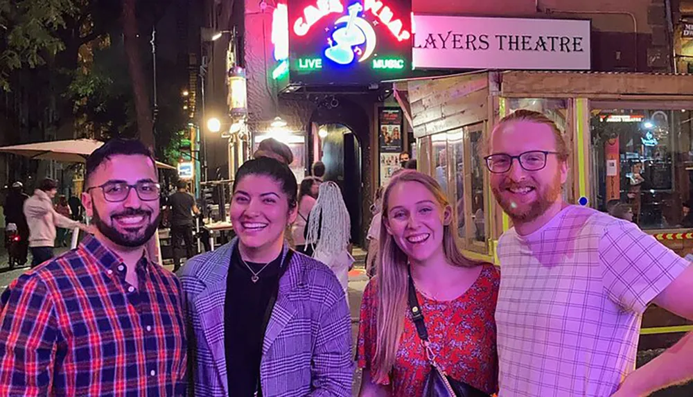 Four smiling individuals are posing for a photo in front of a venue advertising live music with the neon sign Casbah located above The Players Theatre