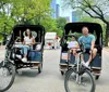 Two people are enjoying a ride in pedicabs through a park with tall buildings in the background