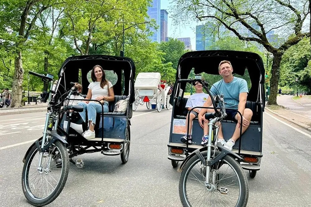 Two people are enjoying a ride in pedicabs through a park with tall buildings in the background