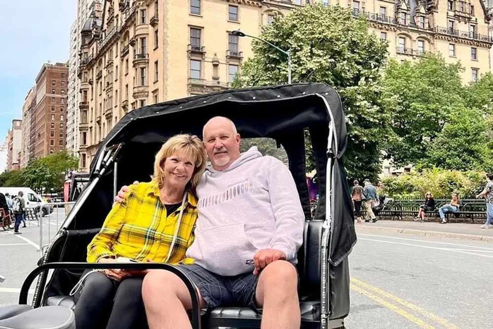 A smiling man and woman are sitting in a pedicab with city buildings and trees in the background