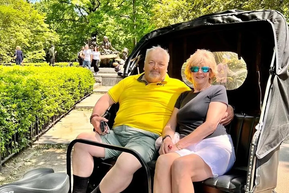 A smiling couple is seated comfortably in a pedicab enjoying a sunny day in a park with green foliage and a statue in the background