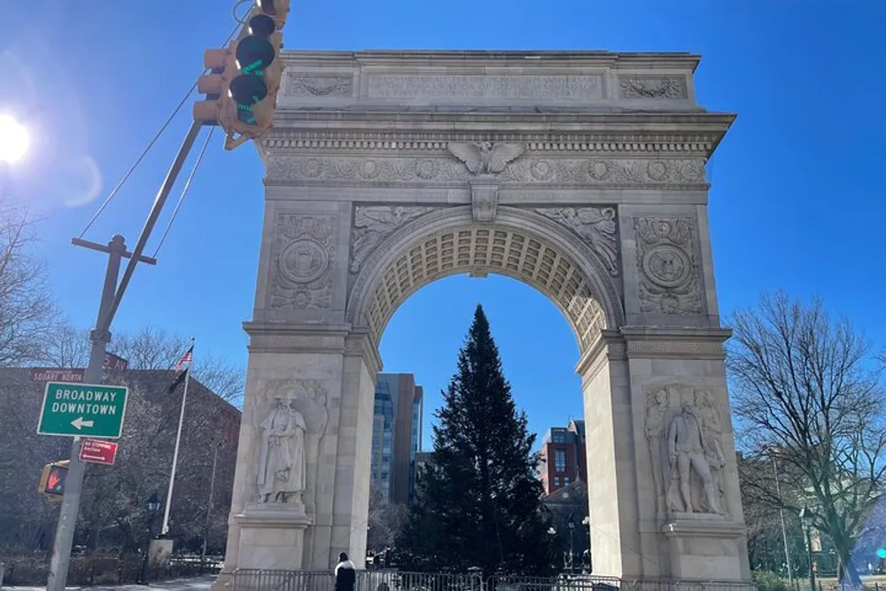The image shows the Washington Square Arch on a sunny day with a clear blue sky a traffic light showing green and street signs for Broadway Downtown