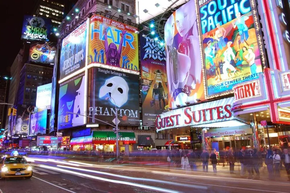 The image shows a bustling Times Square at night with bright billboards for Broadway shows glowing neon lights and the motion blur of moving vehicles and pedestrians