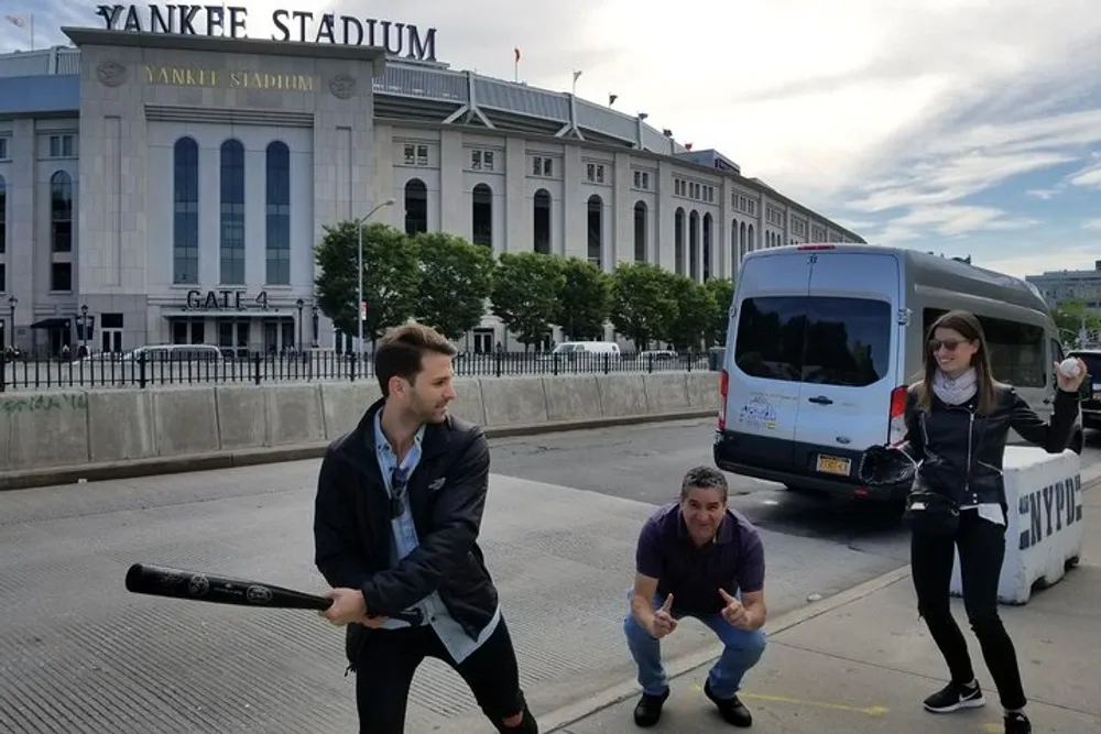 Three people are playfully mimicking baseball actions in front of the Yankee Stadium