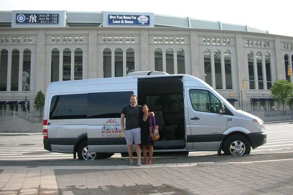 Two individuals are standing and smiling in front of a parked travel van with a large stadium structure visible in the background