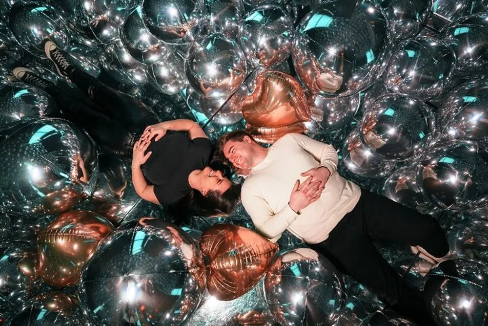 Two people are lying amidst a sea of silvery and golden balloons creating an immersive reflective environment