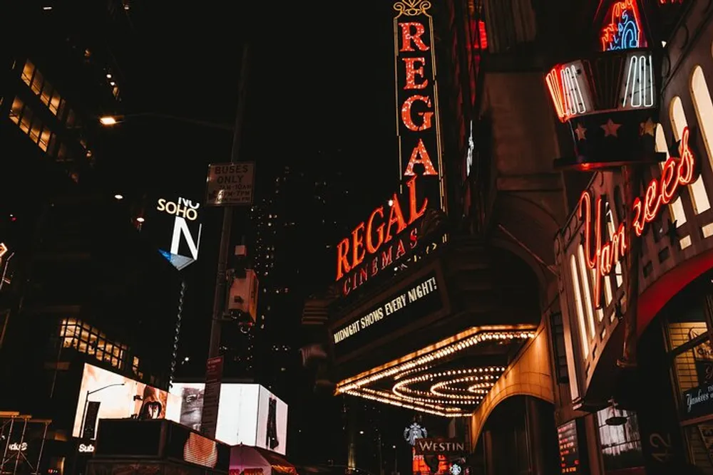 The image captures a vibrant urban night scene with illuminated signage of the REGAL cinemas and adjacent businesses creating an inviting atmosphere along the bustling street