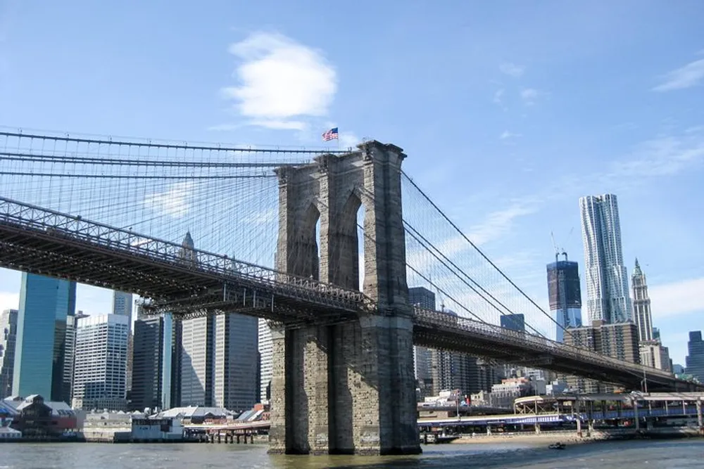 The image shows the Brooklyn Bridge spanning across the East River with the Manhattan skyline in the background on a clear day