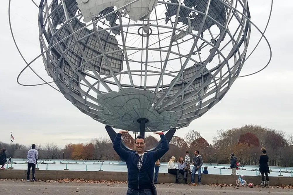 A person is posing with their hands up as if holding the Unisphere a large globe structure at Flushing Meadows-Corona Park in New York