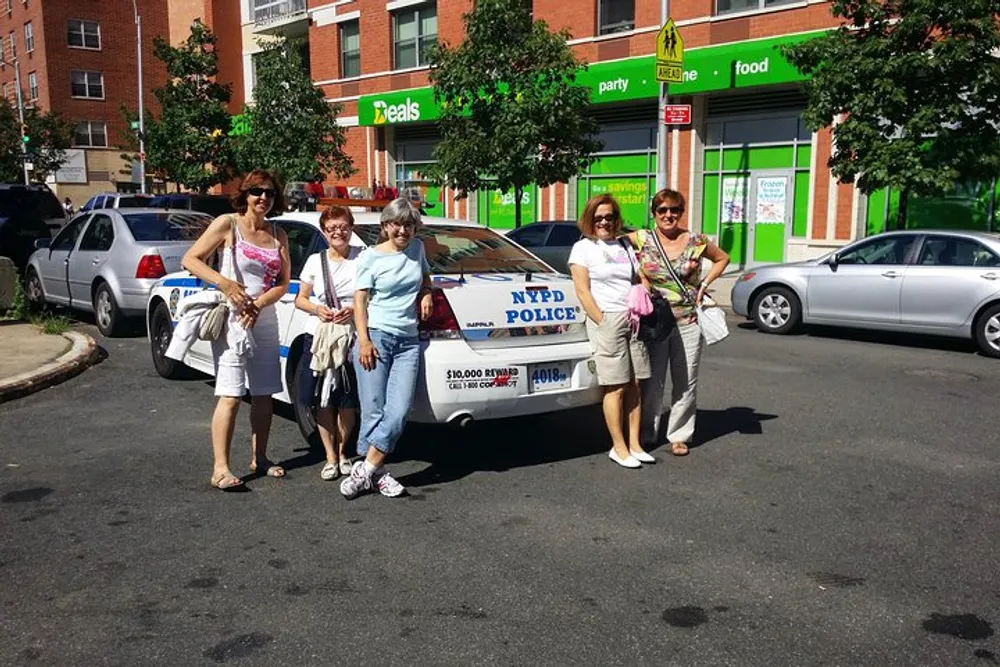 A group of women is posing for a photo on a sunny street next to an NYPD police car