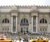 This is a busy day outside the entrance of the Metropolitan Museum of Art in New York City highlighted by bustling pedestrians and classic yellow taxis
