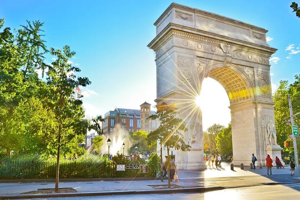 The image portrays a sunny day at Washington Square Park with the suns rays beaming through the arch highlighting people and greenery in a vibrant urban setting
