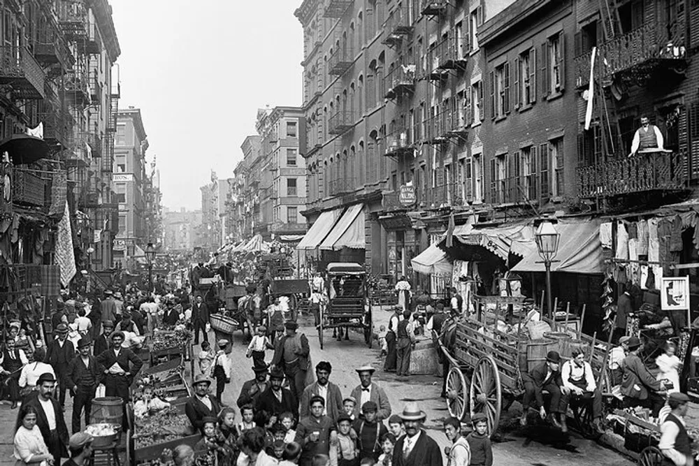 This black-and-white photograph depicts a bustling street scene from an earlier era with people mingling horse-drawn vehicles and market stalls lining the road showcasing a snapshot of urban life from the past