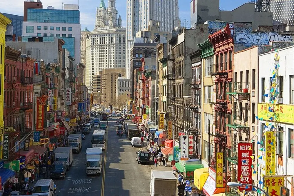 The image captures a busy street lined with colorful buildings and signs indicative of a vibrant Chinatown with cars and pedestrians against a backdrop of skyscrapers under a clear sky