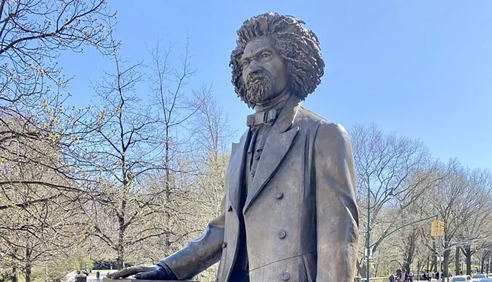 The image shows a bronze statue of a man with distinctive hair and beard set against a backdrop of bare trees and a clear blue sky