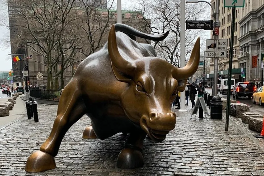 The image shows the iconic Charging Bull statue on a cobblestone street with pedestrians and traffic in the background presumably in the Financial District of Manhattan