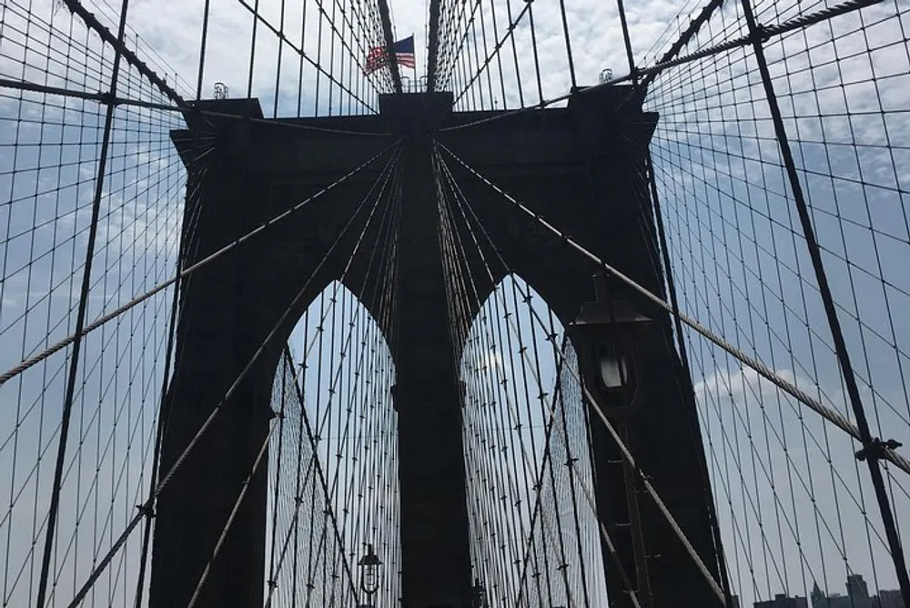 This is a photo taken from the pedestrian walkway of the Brooklyn Bridge showcasing its iconic web of cables and one of its Gothic-style stone towers under a partly cloudy sky
