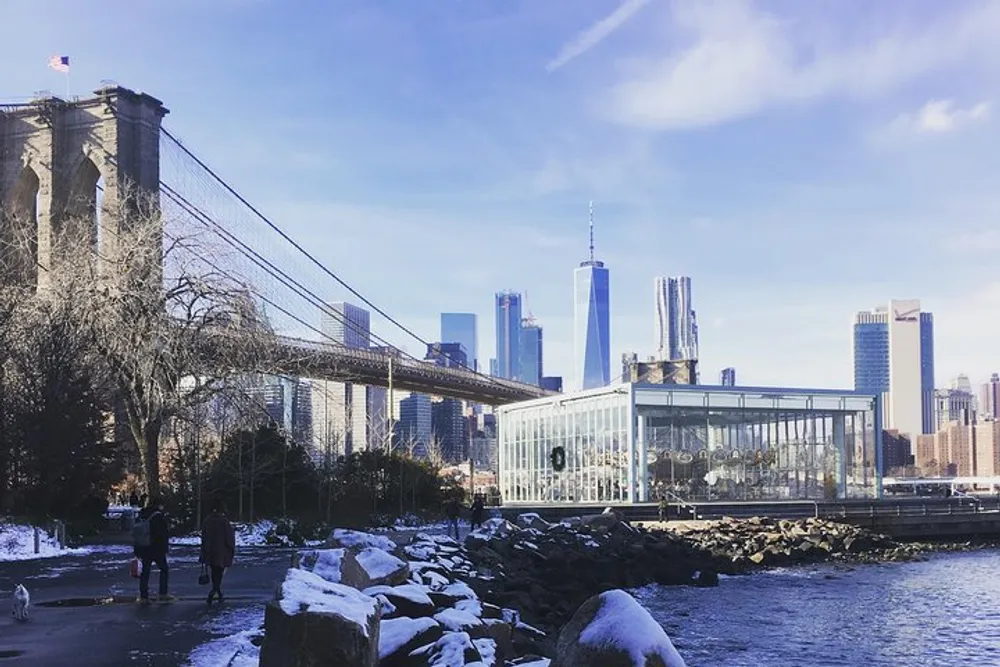 The image shows a snow-dusted park with people near the Brooklyn Bridge with the Manhattan skyline featuring the One World Trade Center in the background under a clear blue sky