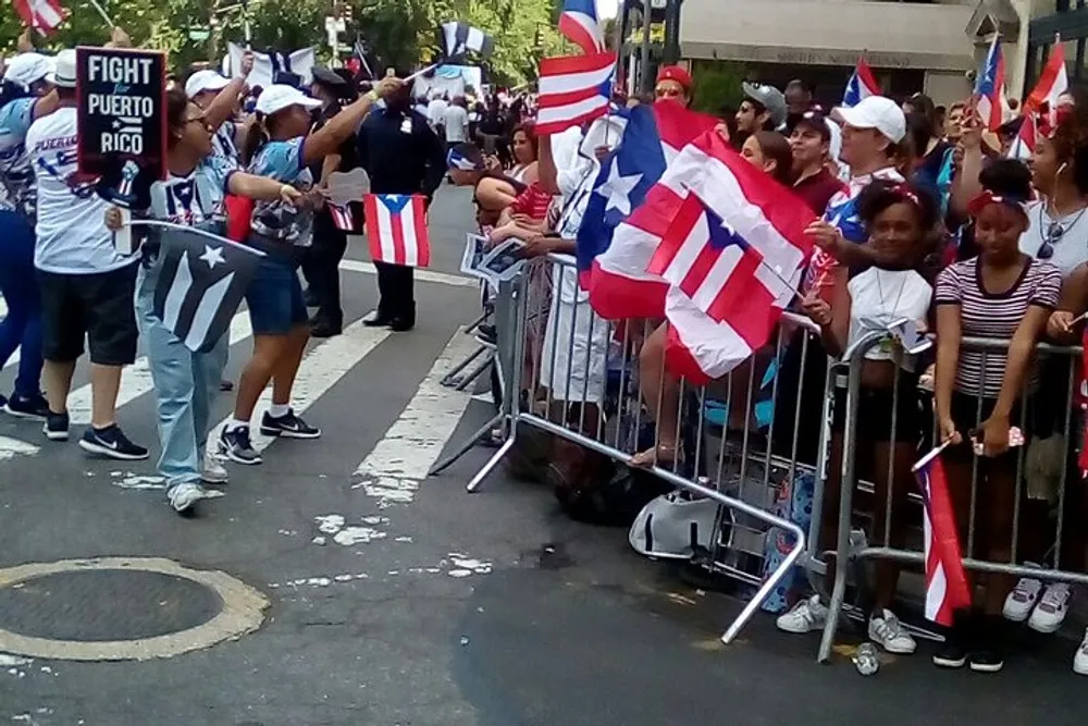 The image shows a group of people some waving Puerto Rican flags standing behind barricades on a city street suggesting a public event or celebration related to Puerto Rico