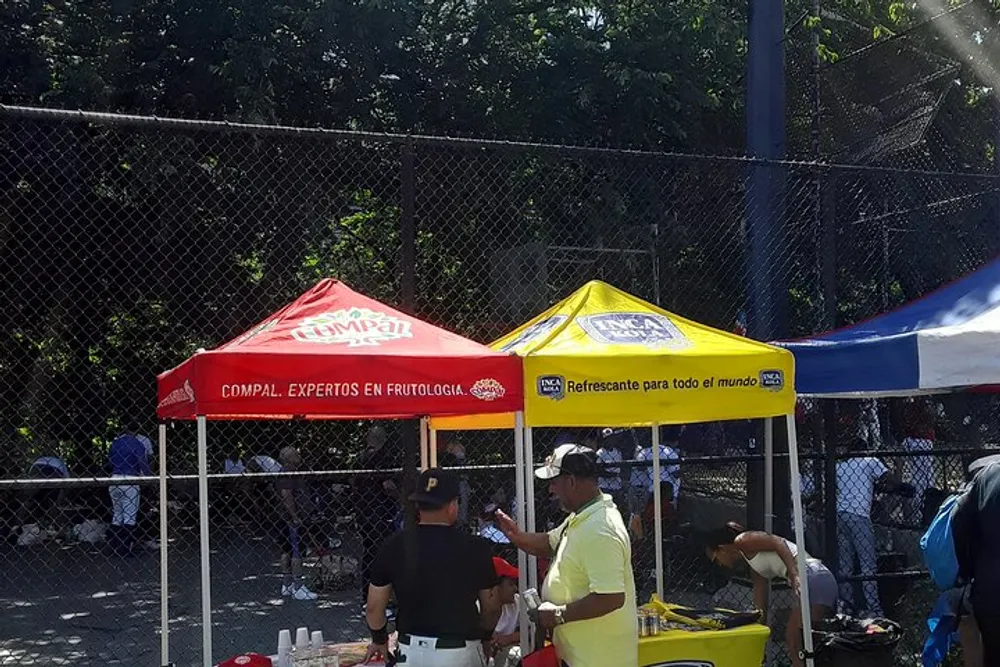 The image shows a sunny outdoor setting with people gathered under branded red and yellow tents possibly at a sports event or community gathering