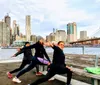 Three people are performing yoga poses on a waterfront with a city skyline and a bridge in the background