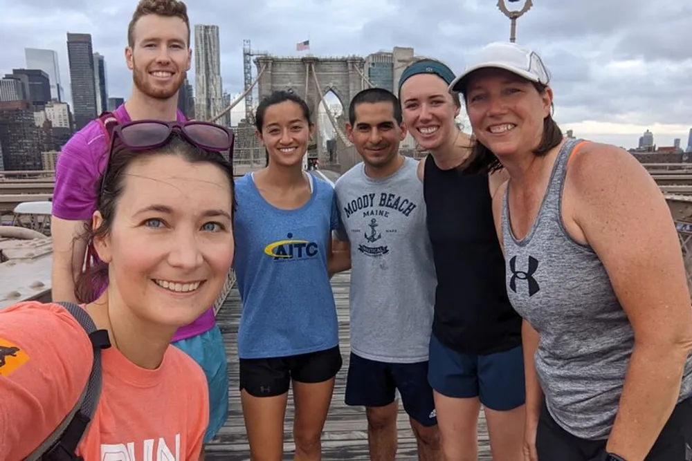 A group of six people are posing for a selfie with the Brooklyn Bridge in the background looking happy and possibly after a run given their sportswear
