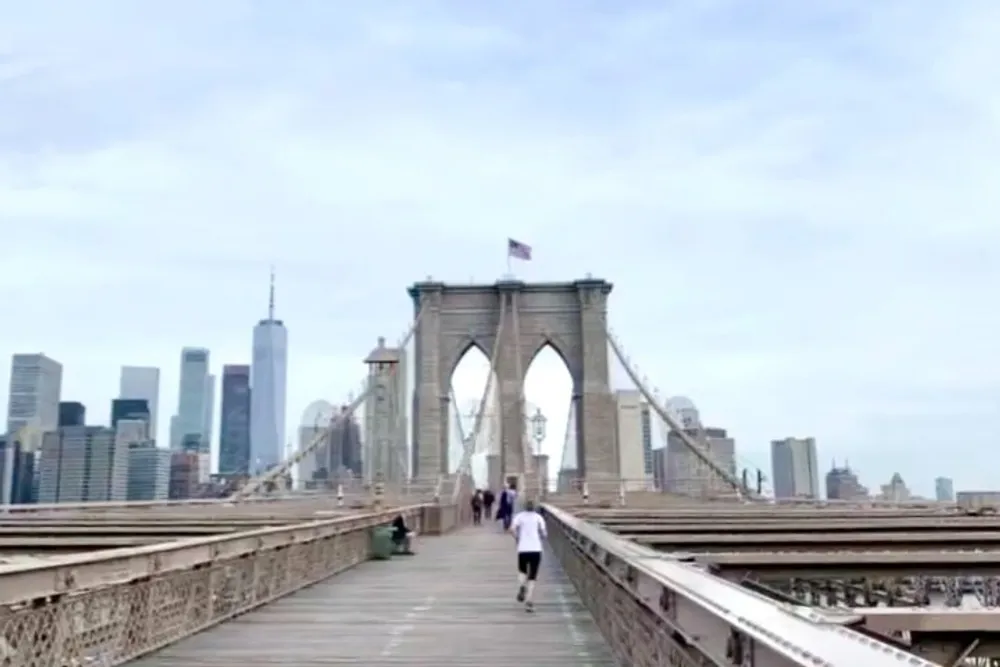 The image shows a person jogging on the pedestrian walkway of the Brooklyn Bridge with the New York City skyline in the background