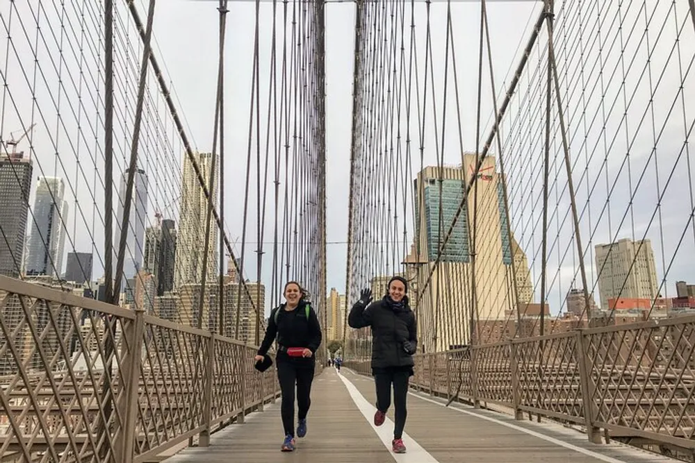 Two people are running on the pedestrian walkway of the Brooklyn Bridge with the New York City skyline in the background