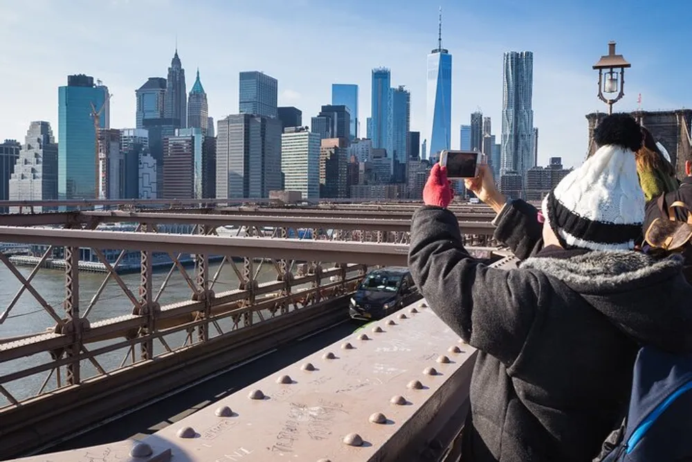 A person is taking a photo with their smartphone of the New York City skyline from a bridge on a clear day