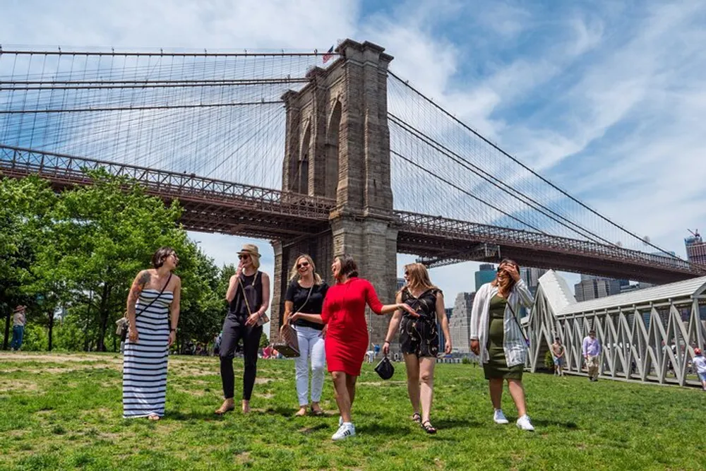 A group of people is enjoying a sunny day in a grassy area with the Brooklyn Bridge in the background