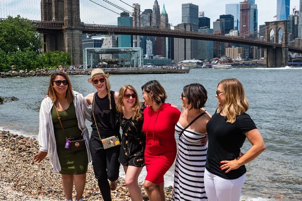 A group of five women are smiling and posing together on a rocky riverbank with the Brooklyn Bridge and Manhattan skyline in the background