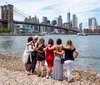 A group of people is enjoying a sunny day in a grassy area with the Brooklyn Bridge in the background