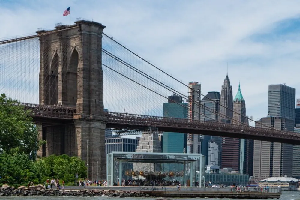The image shows the Brooklyn Bridge with the New York City skyline in the background an American flag atop the bridges tower and a carousel enclosed in glass near the waterfront