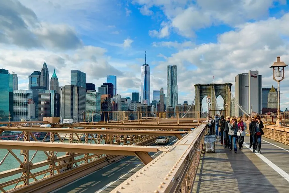 Pedestrians are walking along the Brooklyn Bridge with the New York City skyline including the One World Trade Center in the background under a partly cloudy sky
