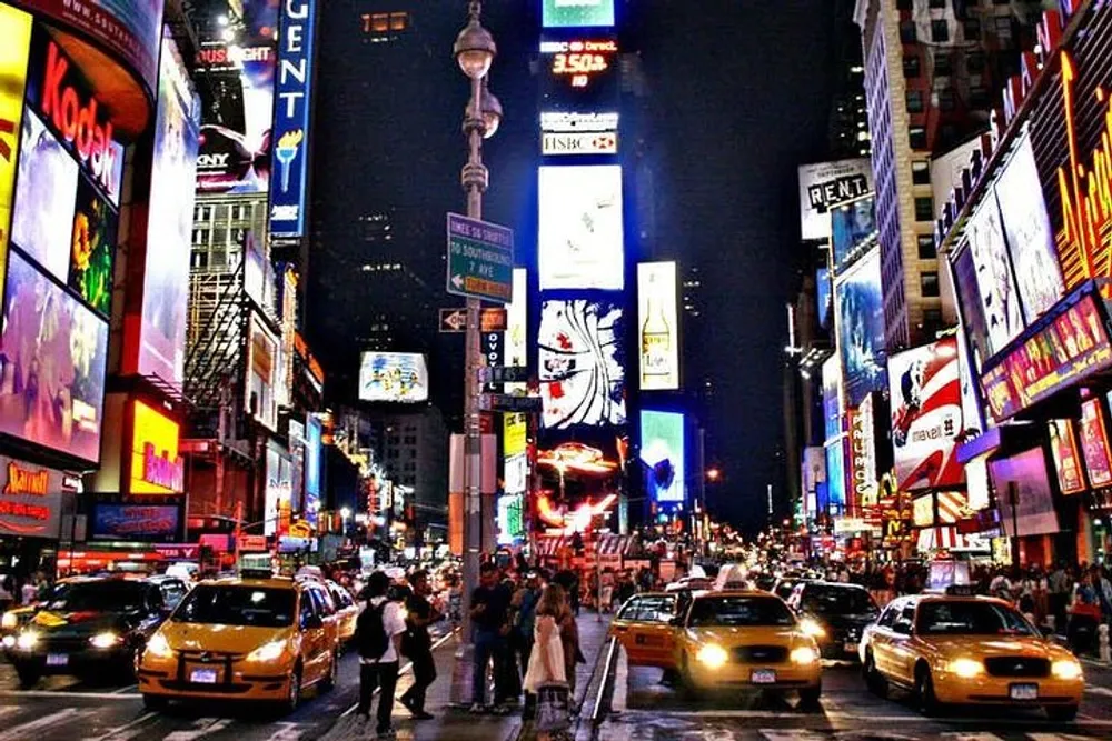 This image captures a bustling Times Square at night with bright billboards illuminating the scene and yellow taxis dominating the street