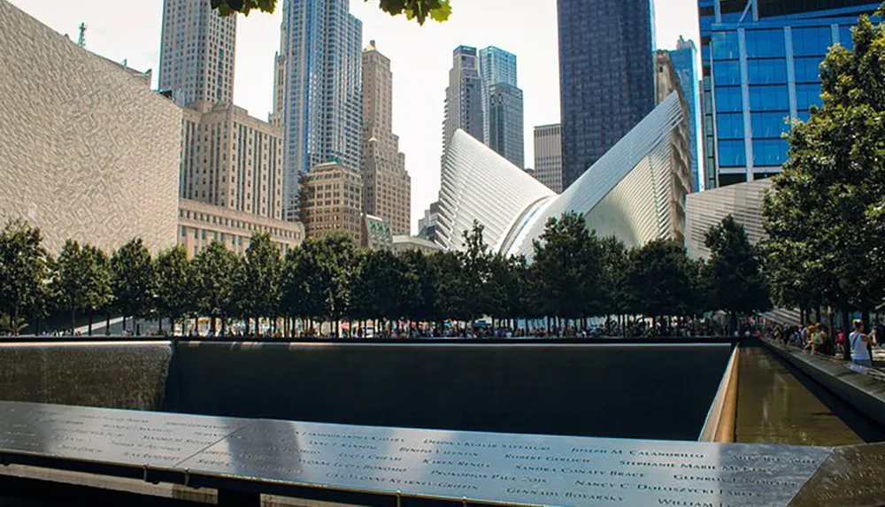 The image shows the National September 11 Memorial with the names of the victims inscribed around the reflecting pools and the Oculus structure in the background amidst the New York City skyline