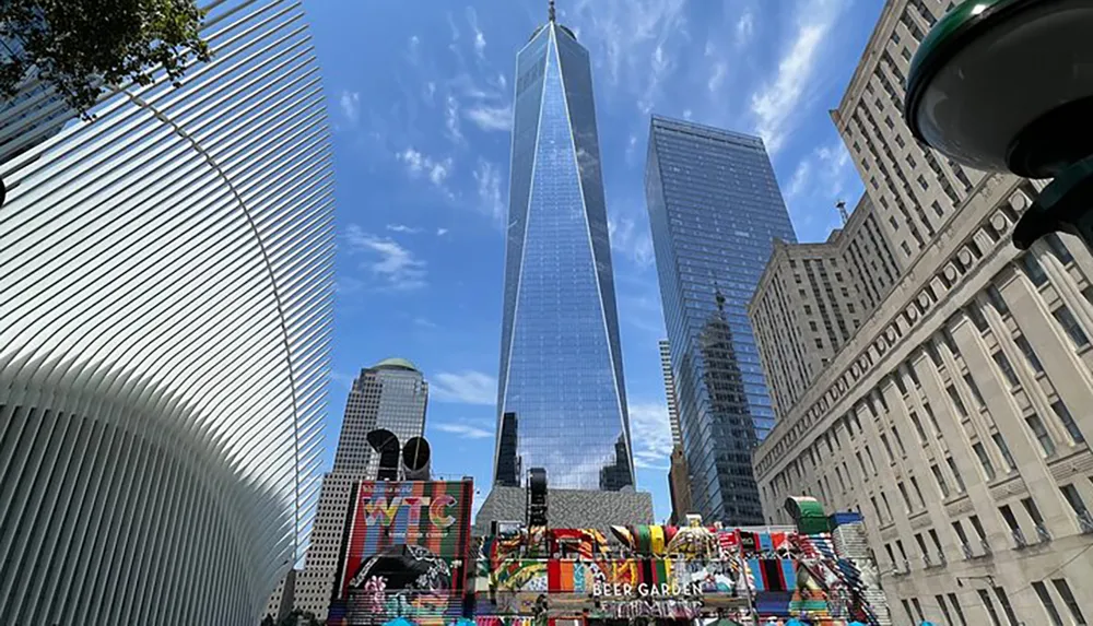 The image showcases a vibrant urban scene with the iconic One World Trade Center towering in the background adjacent to distinctive architecture and a colorful mural under a clear blue sky