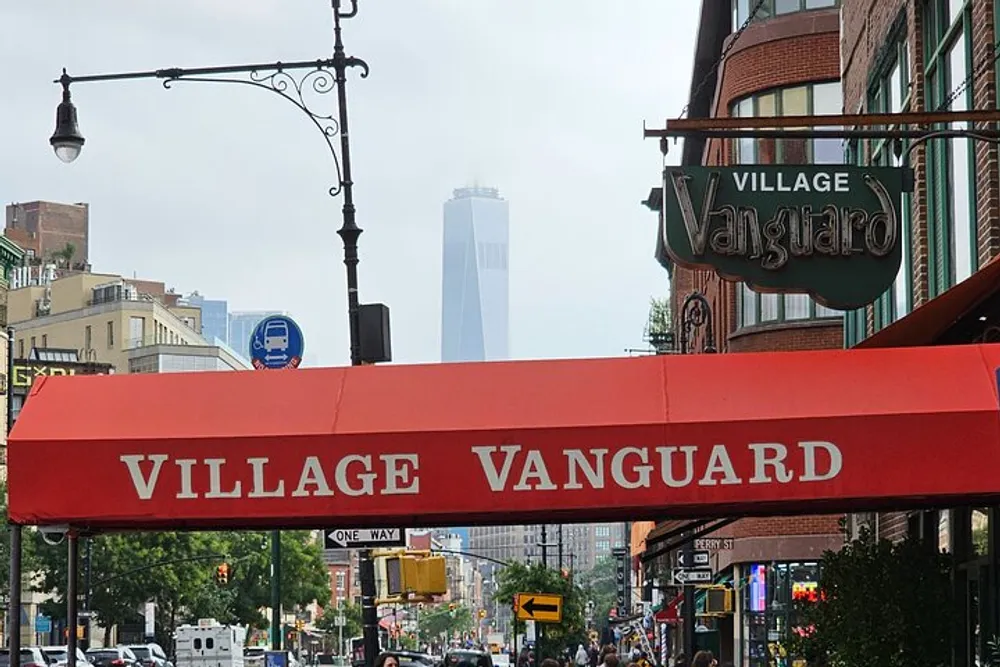 The image captures a daytime street scene with the VILLAGE VANGUARD canopy in the foreground and the silhouette of a tall building in the distance indicative of an urban setting likely in New York City