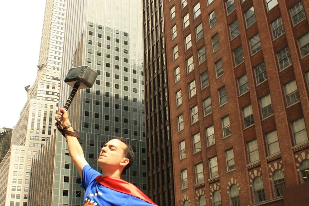A person dressed in a Superman costume is holding a hammer prop aloft amidst city buildings