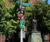 The image features a collection of street signs at an intersection with a statue in the background surrounded by lush green trees