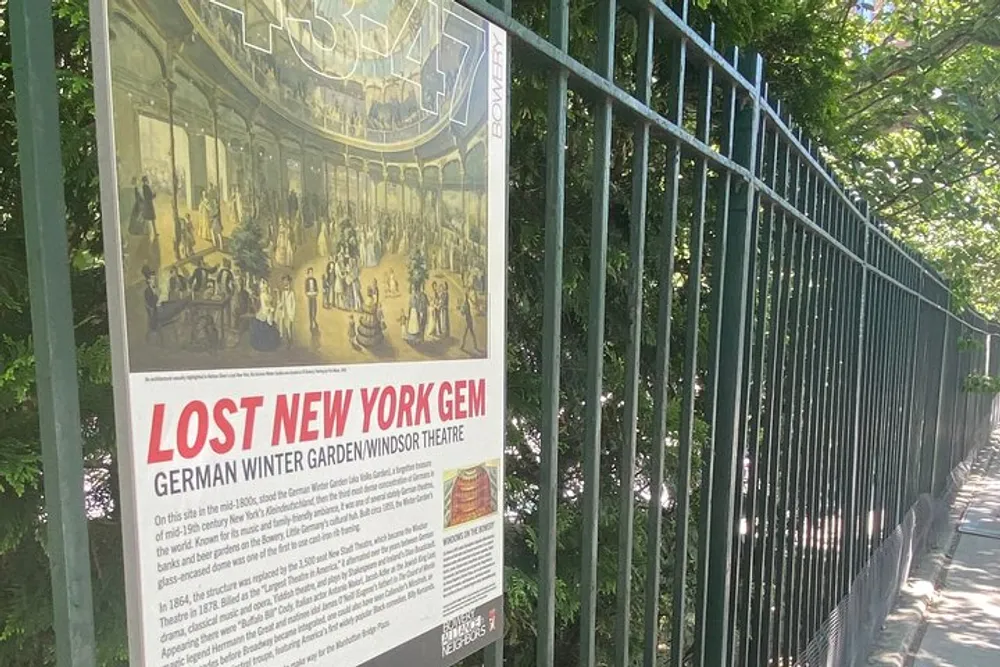 The image shows an information plaque titled LOST NEW YORK GEM German Winter GardenWindsor Theatre mounted on a metal fence along a sidewalk with greenery in the background