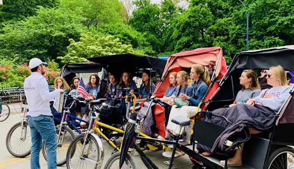 A tour guide is speaking to a group of people seated in pedal-powered rickshaws in a park setting