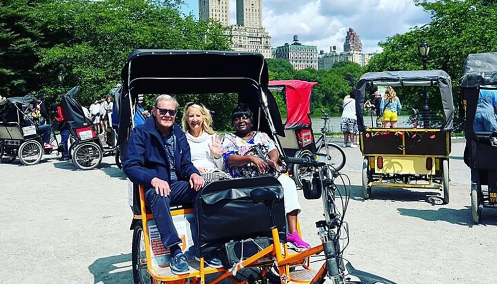 Three people are sitting in a pedal-powered rickshaw enjoying a sunny day in a park with other rickshaws and visitors in the background