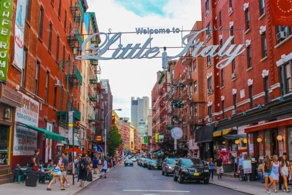 The image shows a vibrant street view with people walking shops lining the road vehicles parked and moving under a sign that reads Welcome to Little Italy