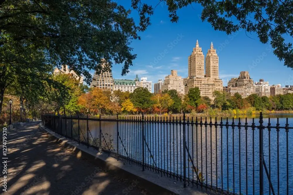 The image shows a peaceful autumn scene with a view of the water and colorful trees in a park foreground complemented by a backdrop of towering skyscrapers under a clear blue sky